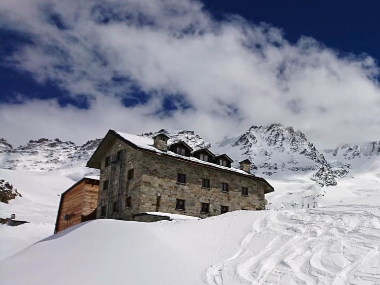 Winteraufnahme des Rifugio Chabod umgeben von tiefem Schnee und zahlreichen Skispuren im Pulverschnee.
