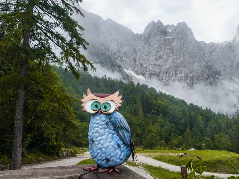 Aussichtsterrasse am Rifugio Luigi Zacchi mit blauer Eulen-Skulptur und Blick über die bewölkten Täler der Alpen.