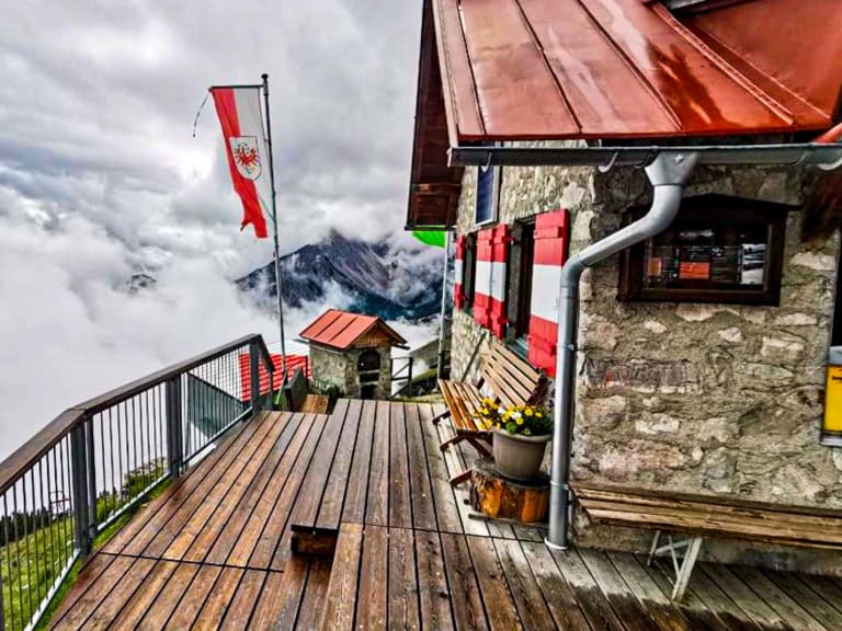 Holzterrasse der Bettelwurfhütte mit Blick über die Wolkendecke auf schneebedeckte Karwendel-Gipfel.