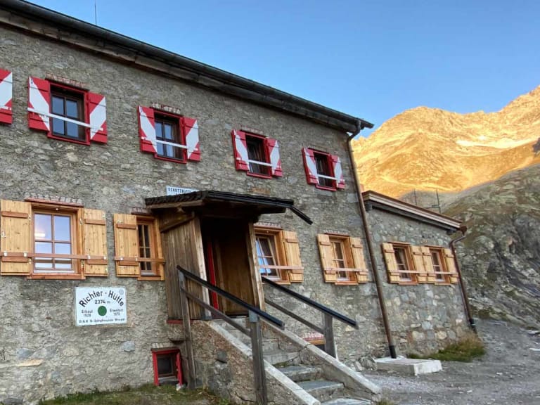 Außenansicht der Richterhütte aus Naturstein mit rot-weißen Fensterläden vor Bergkulisse.
