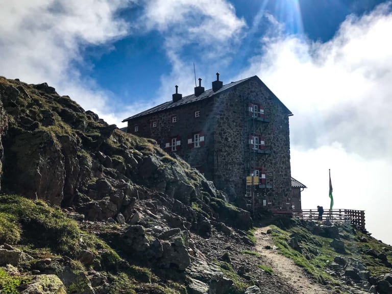 Die Breslauer Hütte aus Naturstein auf felsigem Bergkamm mit grün-weißen Fensterläden und Fahne in den Ötztaler Alpen.
