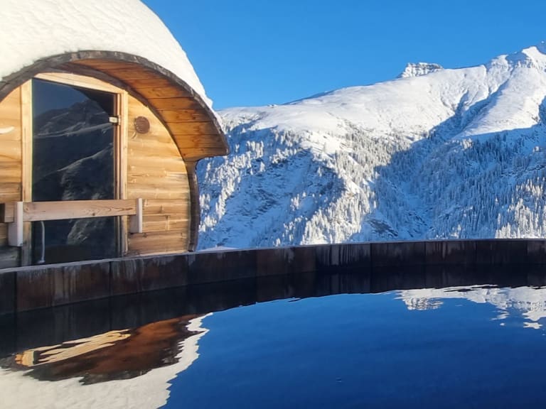 Außenpool mit Sauna-Tonne am Hotel Camana vor verschneiter Bergkulisse der Adula-Alpen bei Sonnenschein.