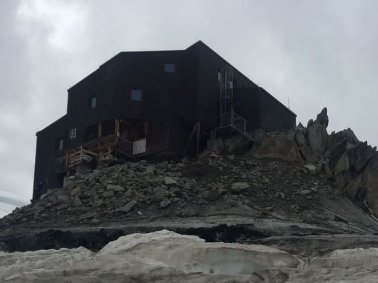 Das Refuge des Cosmiques steht auf einem Felsgrat zwischen Firnfeldern bei bewölktem Himmel.