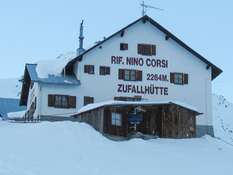 Verschneite Zufallhütte mit weißem Hauptgebäude und Holzanbau in winterlicher Hochgebirgslandschaft.