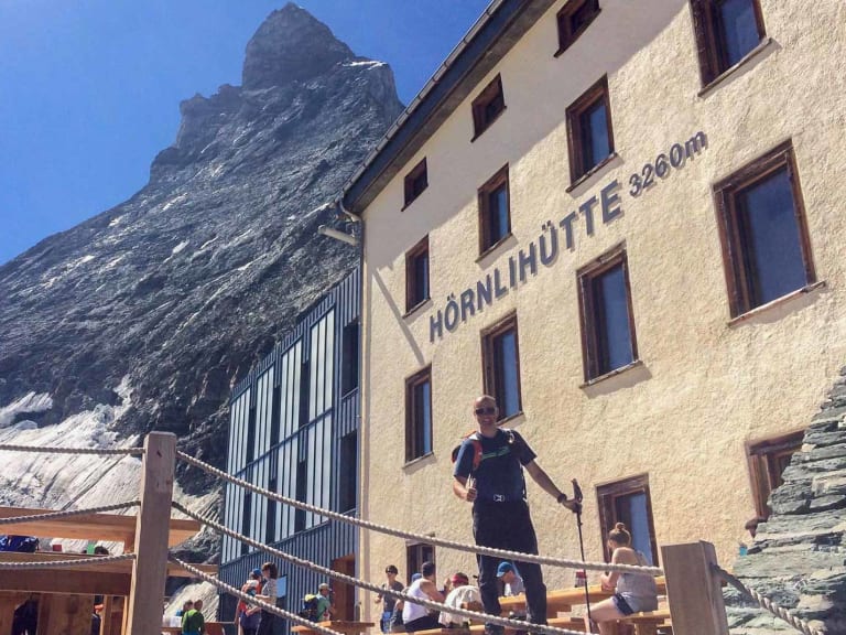 Die Hörnlihütte bei klarem Himmel mit Wanderern auf der Terrasse und steilem Felsgipfel dahinter.