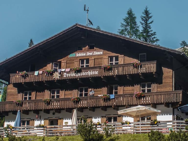 Blick auf die Drei Schuster Hütte mit Holzfassade, Balkonen und Sonnenterrasse vor steilen Felswänden.