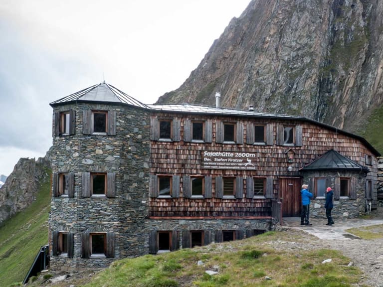 Zwei Wanderer stehen vor der Sajathütte, einer Berghütte aus Naturstein und Holz auf 2.600 m Höhe.
