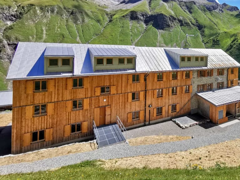 Luftbild der Kemptner Hütte mit mehreren Holzgebäuden und Solarpaneelen auf grüner Almwiese.