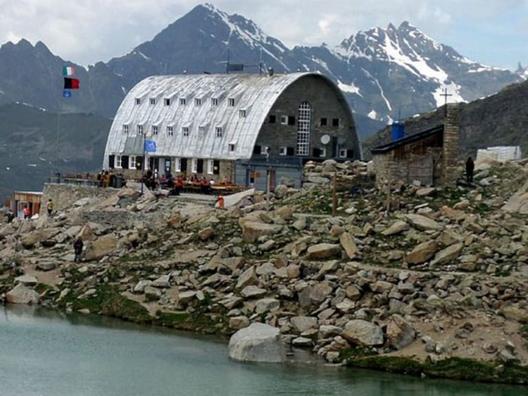Wanderer vor dem Rifugio Vittorio Emanuele II mit charakteristischem Tonnendach und kleinem Bergsee im Vordergrund.
