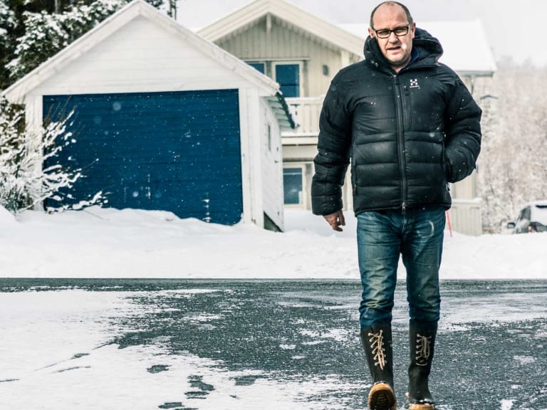 Mann in schwarzer Winterjacke steht mit Hund bei Koppangen Brygger während starkem Schneefall vor weißen Holzhäusern.