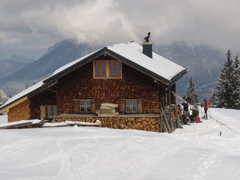 Winteraufnahme der Stuibenhütte mit Gästen auf der verschneiten Terrasse bei bewölktem Wetter.