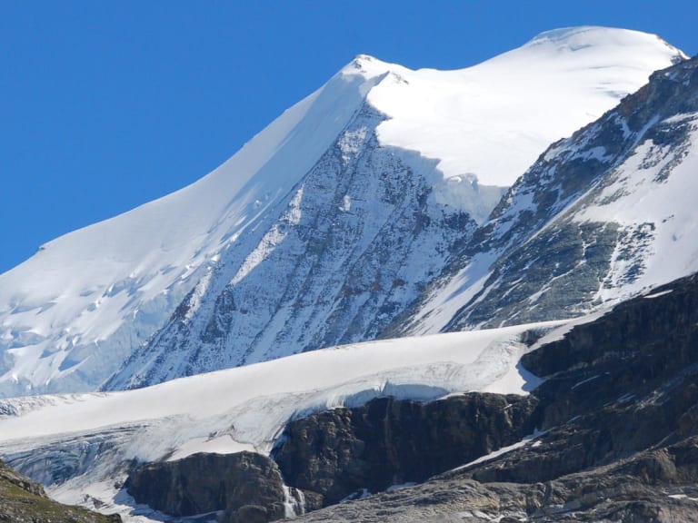 Die Turtmannhütte steht auf einem grasbewachsenen Hang vor verschneiten Gipfeln und einem Gletscher mit Spalten.