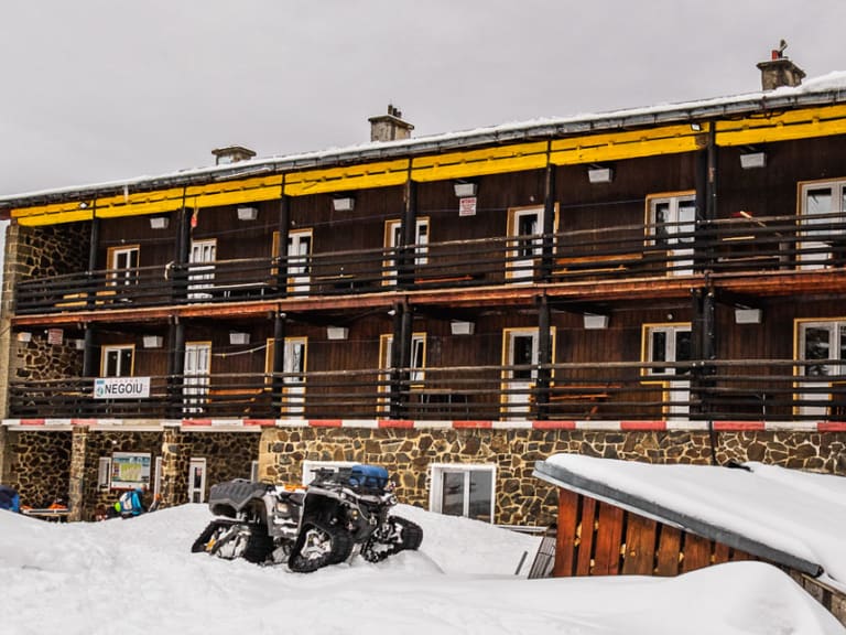 Dreistöckiges Hauptgebäude der Cabana Negoiu mit gelbem Balkon und dicke Schneeschicht auf dem Dach.