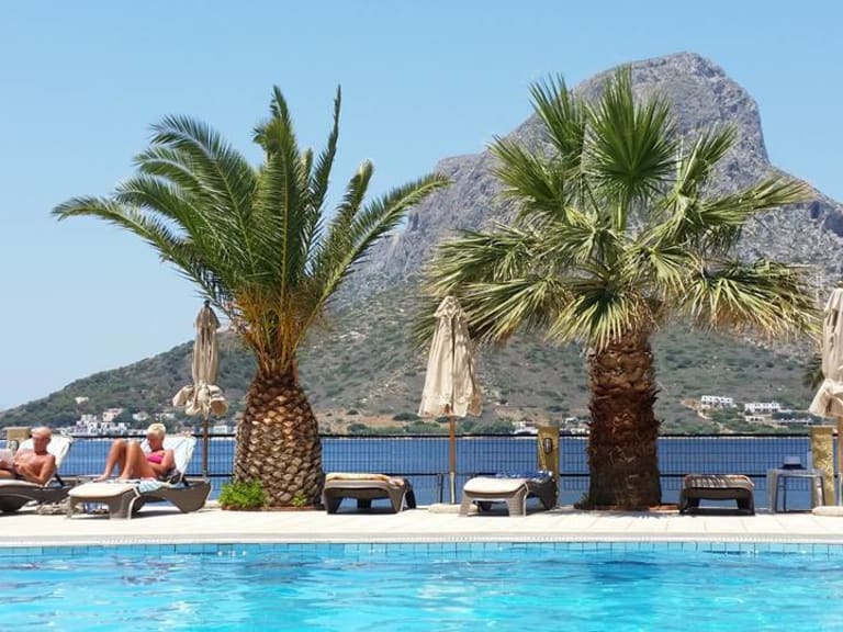 Poolbereich des Hotel Plaza Kalymnos mit Palmen, Liegestühlen und Blick auf die blaue Meeresbucht.