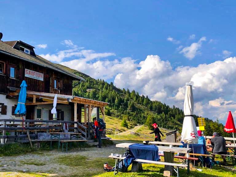 Wanderer versammeln sich vor der Galflunalm mit großer Sonnenterrasse und bunten Schirmen auf grüner Almwiese.