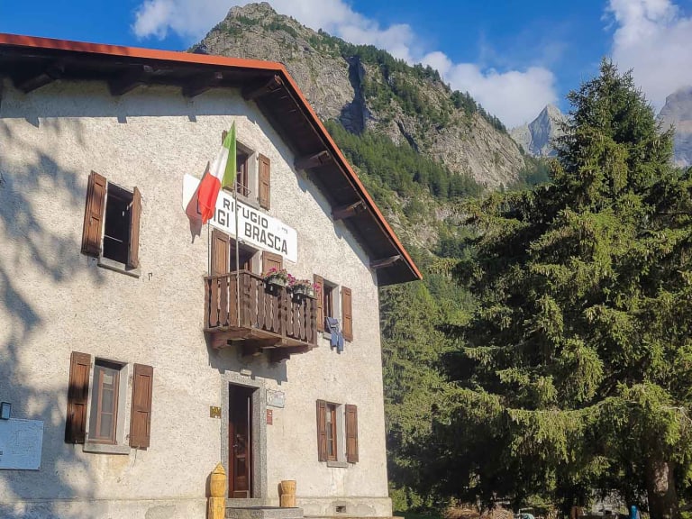 Außenansicht des Rifugio Brasca, einer zweistöckigen Berghütte mit heller Fassade und roten Ziegeldach in alpiner Umgebu