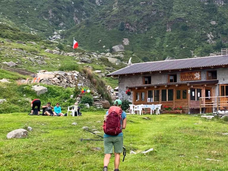 Wanderer mit rotem Rucksack vor dem Rifugio Alpe Baranca auf grüner Almwiese in den Walliser Alpen.