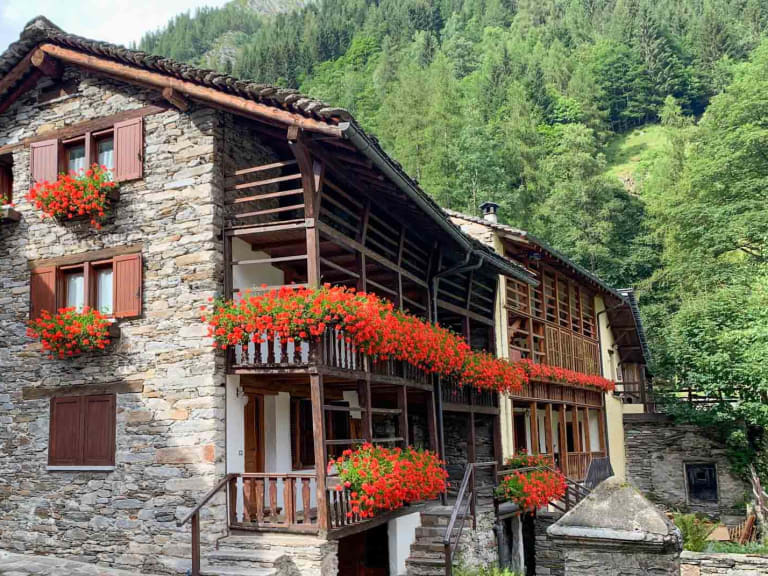 Traditionelle Steinhäuser mit Holzbalkonen und roten Geranien umgeben das Rifugio Brusà in alpiner Landschaft.