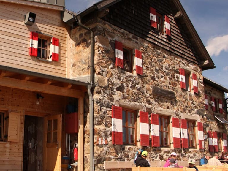 Außenansicht des Schutzhaus Latzfonser Kreuz mit Naturstein-Holz-Konstruktion und sonniger Terrasse auf 2.296 m Höhe.
