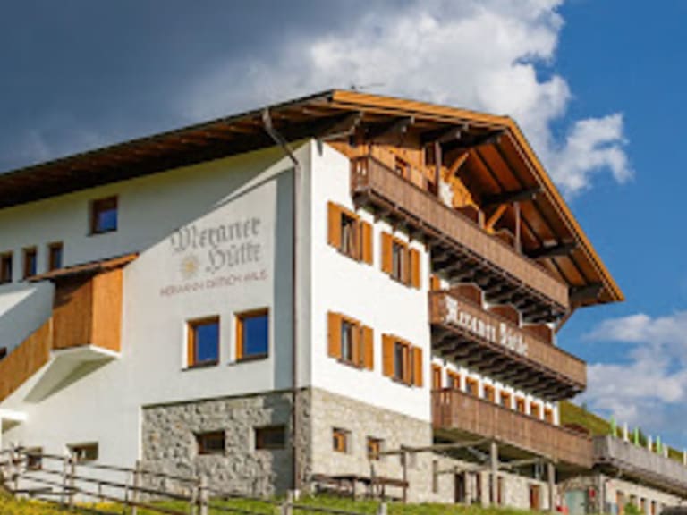 Die Meraner Hütte zeigt sich mit weißer Fassade, Holzbalkonen und traditionellem Satteldach auf grüner Almwiese.