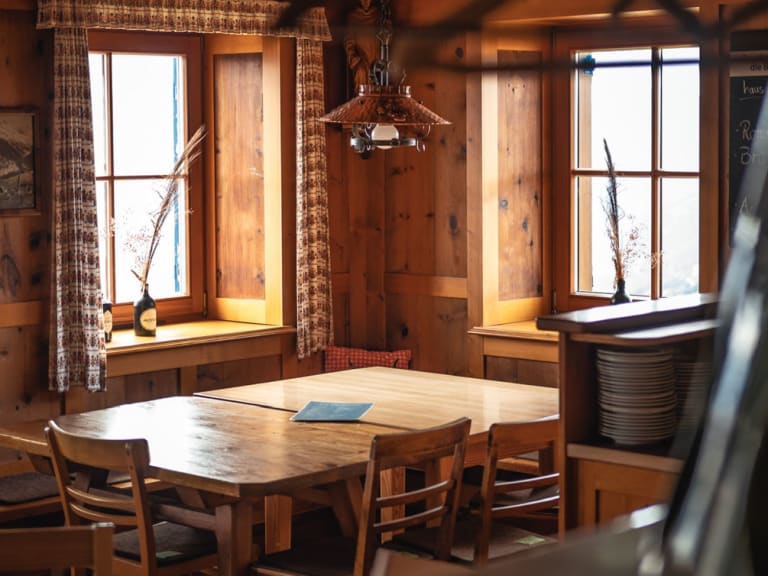 Gemütlicher Gastraum der Leutkircher Hütte mit Holztisch, karierten Vorhängen und Blick auf verschneite Berglandschaft.