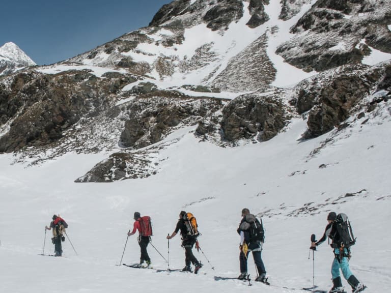 Zehn Skitourengeher steigen durch Tiefschnee zur Cabane de Chanrion auf, die Berghütte ist links im Bild sichtbar.