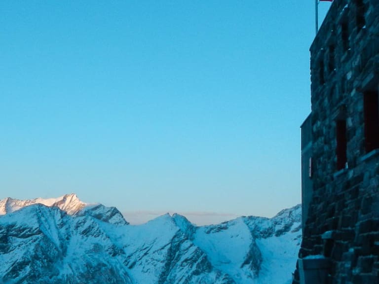 Die Britannia-Hütte aus Naturstein steht verschneit auf einem Felsplateau mit Schweizer Flagge in der Hochgebirgslandschaft.
