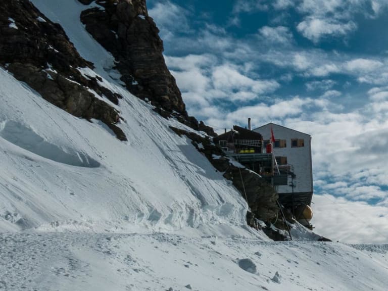 Die Mönchsjochhütte steht auf einem Felssockel umgeben von Schneehängen und schneebedeckten Viertausendern der Berner Alpen.