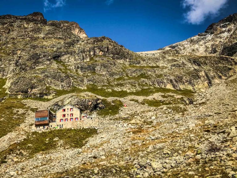 Panoramaaufnahme der Tschiervahütte auf einem Plateau umgeben von Geröllfeldern mit Felswand im Hintergrund der Bernina-Alpen.