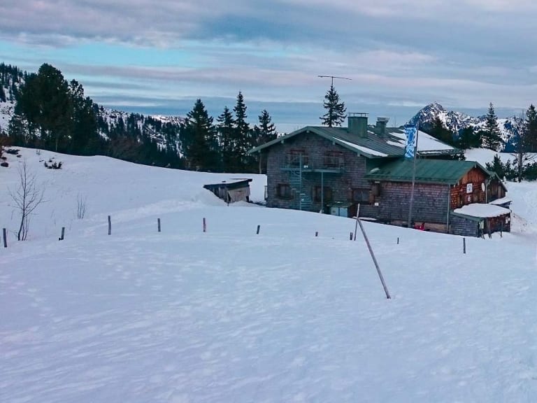 Das Taubensteinhaus steht im Winter auf einem verschneiten Plateau in den Bayerischen Voralpen mit Bergpanorama.