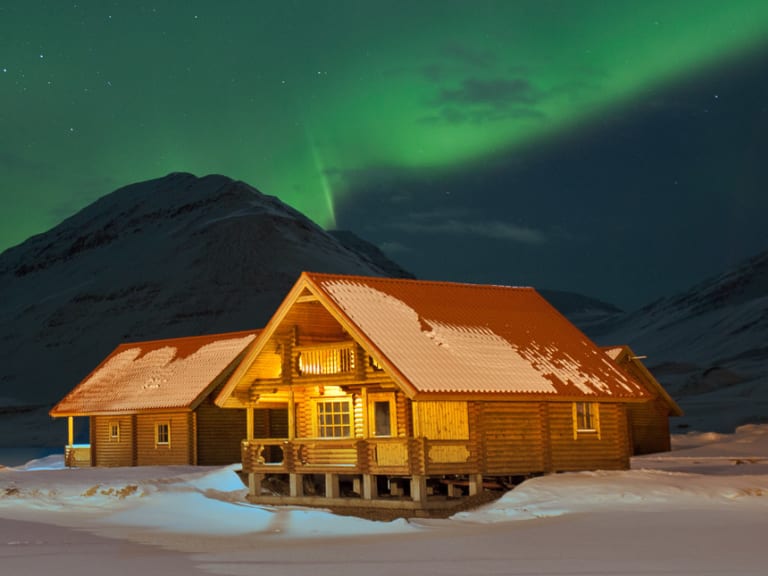 Das beleuchtete Brimnes Hotel and Cabins steht unter grünen Nordlichtern in verschneiter Landschaft Islands.
