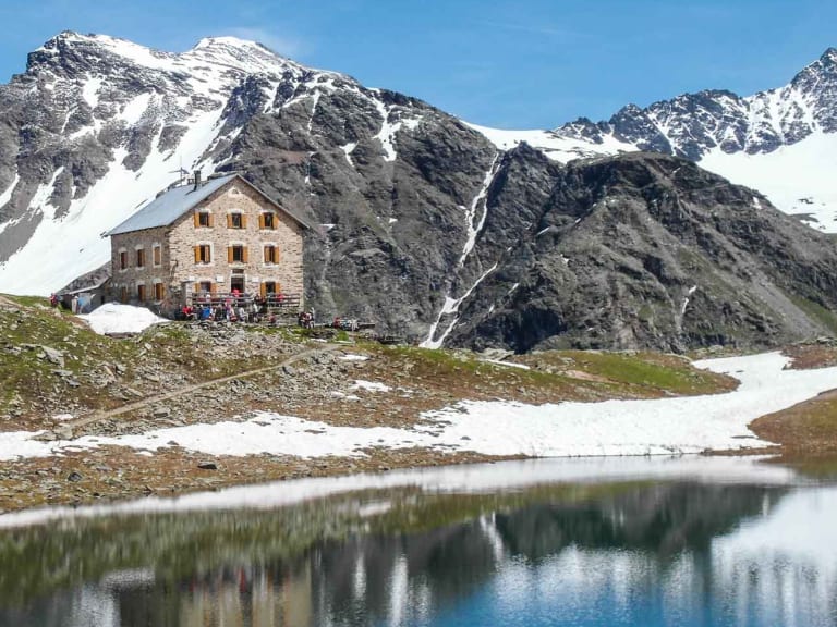 Die Hintergrathütte steht auf einem Plateau oberhalb eines Bergsees, umgeben von schneebedeckten Gipfeln im Ortlergebiet.