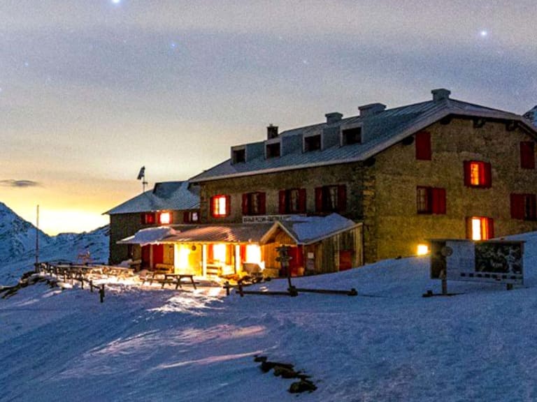 Das Rifugio Pizzini zeigt sich bei Nacht mit warmer Innenbeleuchtung in tiefverschneiter Hochgebirgslandschaft auf 2706 m.