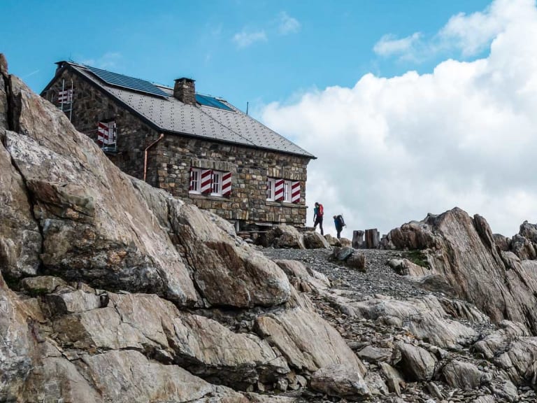Die Tierberglihütte steht auf felsigem Terrain mit zwei Wanderern vor schneebedeckten Gipfeln der Urner Alpen.