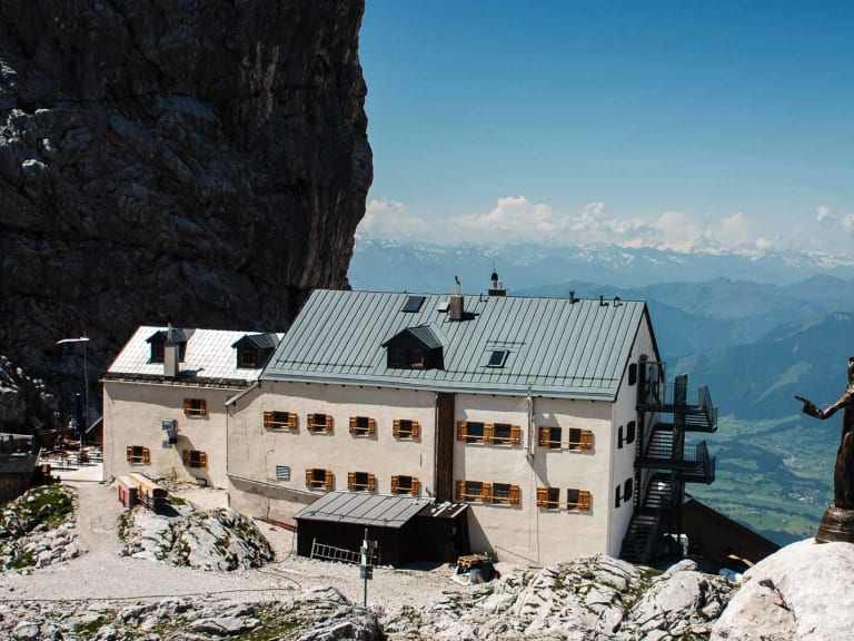 Das Riemannhaus thront auf einem felsigen Plateau in steiler Kalksteinwand mit Wanderer im Vordergrund und weitem Bergpanorama.