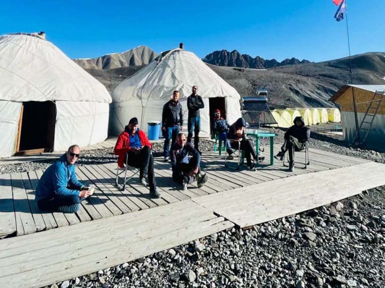 Das Advanced Base Camp ABC Pik Lenin zeigt weiße Jurten auf Holzplattform mit Expeditionsteilnehmern in karger Berglandschaft.