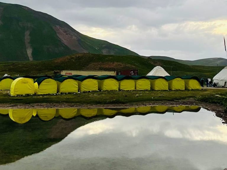 Gelbe Expeditionszelte des Basecamp Pik Lenin spiegeln sich in einem Wassertümpel der kirgisischen Hochebene bei Abendlicht.
