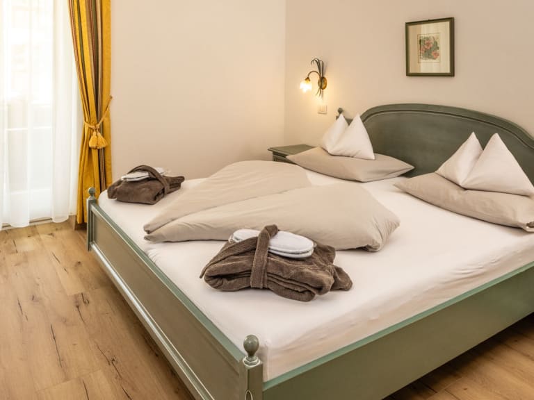 Doppelzimmer im Garni Zimmerhofer Ahrntal mit grünem Holzbett, Glastisch und gelben Vorhängen im traditionellen Alpenstil.