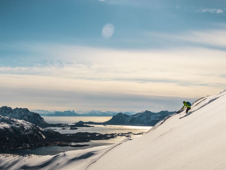 Bergführer Hansi Stöckl in grüner Jacke fährt auf steiler Skipiste bergab mit weitläufiger Fjordlandschaft und verschneiten Bergen im Hintergrund.
