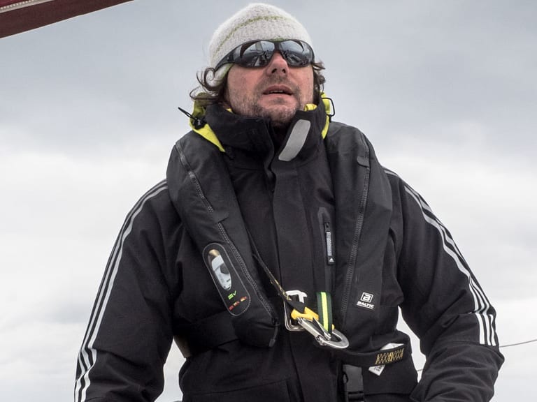 Bergführer Hans Honold in schwarzer Segeljacke und gelber Rettungsweste steuert ein Segelboot bei bewölktem Wetter auf dem Meer.