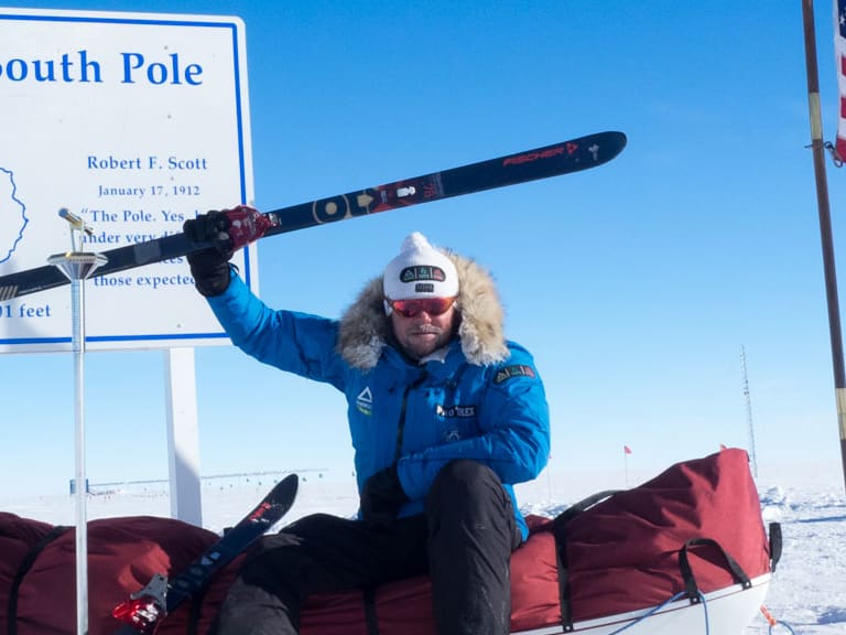 Bergführer Hans Honold sitzt am geografischen Südpol auf rotem Expeditionsschlitten und hält einen Ski über dem Kopf.