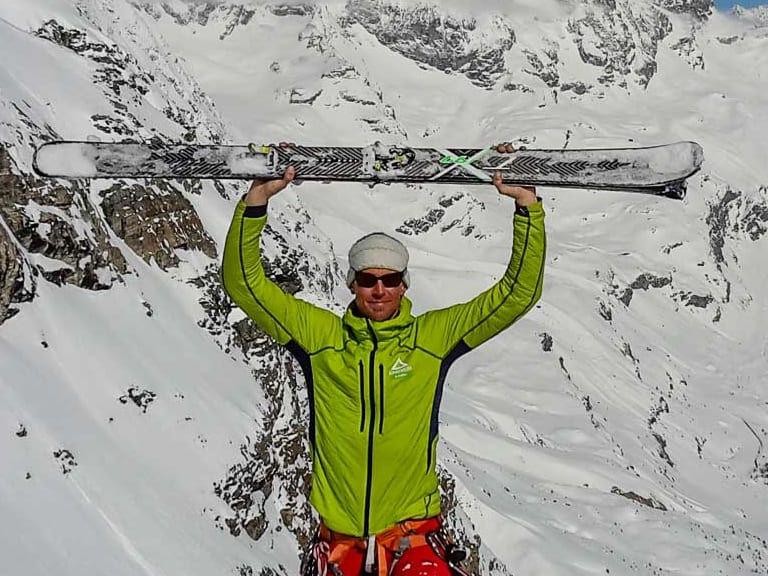 Bergführer Markus Beck hält zwei Skier über dem Kopf vor verschneiter Hochgebirgslandschaft mit pyramidenförmigem Felsgipfel.