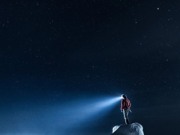 Bergführer Markus Beck steht in roter Winterkleidung auf einem Eisblock unter Sternenhimmel, die Stirnlampe leuchtet durch die Polarnacht.