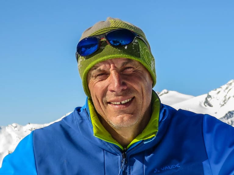 Bergwanderführer Hans Beggel lächelt vor verschneiter Bergkulisse, trägt blaue Jacke mit Alpine Welten Logo und grüne Mütze.