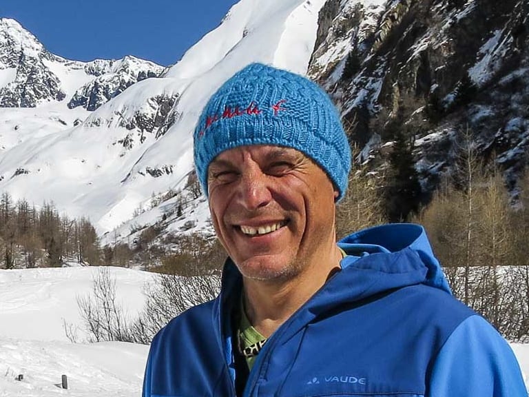 Bergwanderführer Hans Beggel mit blauer Strickmütze und Winterjacke steht lächelnd auf verschneiter Fläche vor tiefverschneitem Tal.
