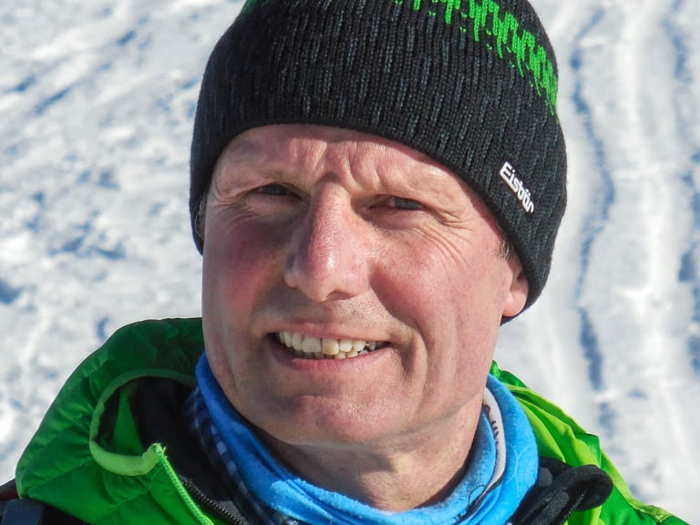 Bergwanderführer Albert Bucher lächelt in die Kamera, trägt schwarze Strickmütze und grüne Winterjacke vor Schneelandschaft.