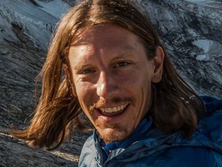 Portrait von Skilehrer Benedikt Fritz mit schulterlangen braunen Haaren und Bart in blauer Softshell-Jacke vor Felswand.