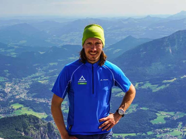 Skilehrer Benedikt Fritz steht lächelnd auf einem Felsgipfel mit grüner Mütze und blauem Funktionsshirt vor Alpenpanorama.