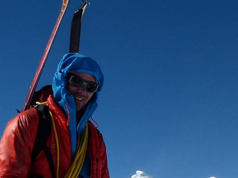 Auf einem schneebedeckten Grat steht Bergführer Bernd Budil in roter Daunenjacke mit Eispickel und gelbem Seil über der Schulter.