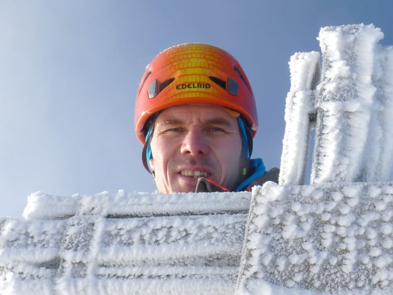 Portrait von Bergführer Bernd Budil mit orangefarbenem Helm am vollständig vereisten Gipfelkreuz bei klarem Himmel.